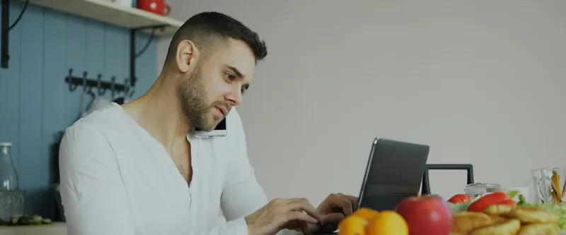 הורה מדבר בטלפון ובודק פרטים במחשב נייד, יושב במטבח